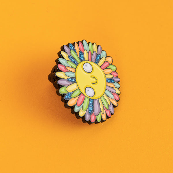 Close-up of Katie Abey's smiling sun enamel pin with glitter accents on its colourful rainbow rays. A bold and quirky accessory full of kindness and fun, perfect for adding a splash of joy to any outfit or bag.