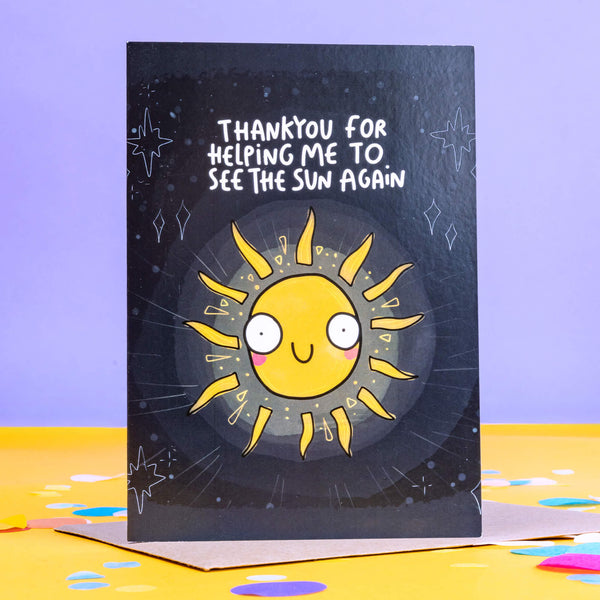 A black greeting card with white twinkling stars and white writing on the front that reads 'thankyou for helping me to see the sun again' and an illustration of a yellow sun with pink cheeks and a smiley face