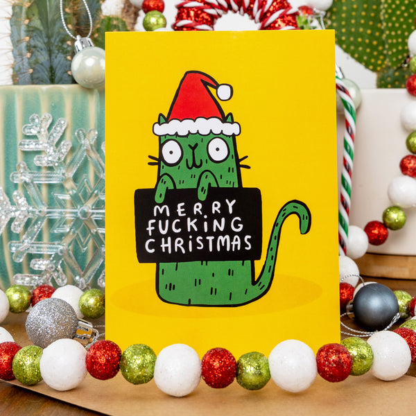 A festive Christmas card from Katie Abey featuring a quirky green cat in a Santa hat, holding a sign that says "Merry Fucking Christmas" on a yellow background. Surrounding the card are holiday decorations, including candy canes, baubles, and a snowflake, creating a vibrant seasonal setting. This card is ideal for people with a taste for sassy humour and non-traditional holiday vibes, embodying Katie Abey's encouragement to embrace your inner weirdness this Christmas.