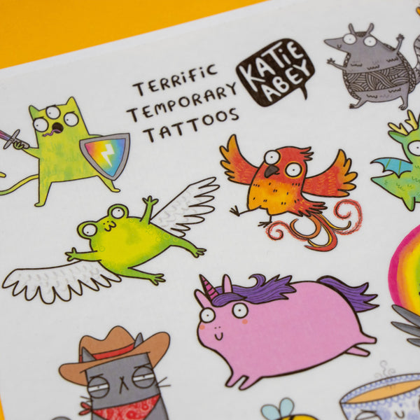 Close up of Katie Abey Terrific Temporary Tattoos sheet showing detailed cartoon illustrations including a flying frog, phoenix bird and unicorn, with bold outlines and vibrant colours on a white background.
