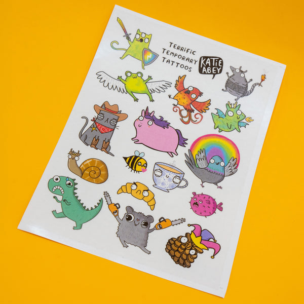 Sheet of colourful Katie Abey Terrific Temporary Tattoos on a bright yellow background, featuring quirky cartoon characters including a unicorn, frog with wings, cowboy cat, rainbow pigeon, dinosaur and snail, in a playful and whimsical illustration style.