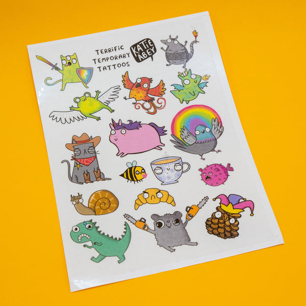 Full sheet of Katie Abey Terrific Temporary Tattoos displayed on a yellow background, packed with cute and quirky illustrated characters including animals, fantasy creatures and humorous designs.