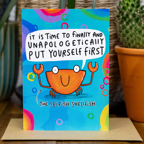 Blue coloured greeting card with rainbow designs and crab illustration on the front with white speech bubble above crabs mouth saying 'it is time to finally and unapologetically put yourself first'. There is also black writing under the crab saying 'The Selfish Shellfish' with an arrow up to the crab.