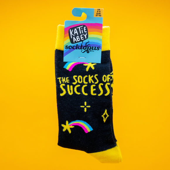 A pair of Katie Abey “The Socks of Success” novelty socks displayed against a bright yellow background. The black socks feature yellow cuffs, toes, and heels, with colourful rainbows, stars, and the uplifting slogan “The Socks of Success”. Fun, quirky design from Katie Abey, the small UK illustrator known for weird, joyful artwork.
