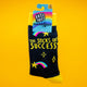 A pair of Katie Abey “The Socks of Success” novelty socks displayed against a bright yellow background. The black socks feature yellow cuffs, toes, and heels, with colourful rainbows, stars, and the uplifting slogan “The Socks of Success”. Fun, quirky design from Katie Abey, the small UK illustrator known for weird, joyful artwork.