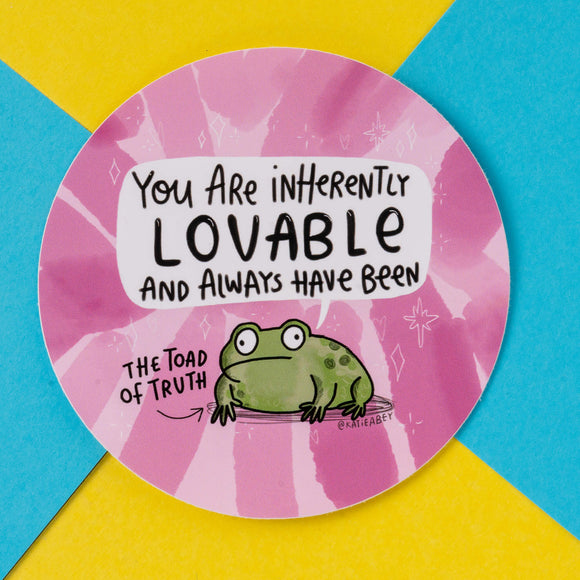 A round sticker featuring a cute, green toad with wide eyes on a pink tie-dye background. The text reads, "You are inherently lovable and always have been," with a label pointing to the toad saying, "The Toad of Truth." This sticker is by Katie Abey, an illustrator known for encouraging people to embrace their weirdness. The background includes geometric patterns, making the design playful and vibrant.