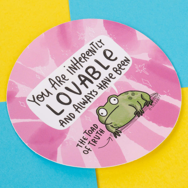 A circular sticker showcasing a green, cartoon toad on a pink tie-dye background with white star accents. The text states, "You are inherently lovable and always have been," while a small label next to the toad identifies it as "The Toad of Truth." This artwork is by Katie Abey, a small business illustrator who promotes self-love and embracing individuality.