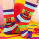 A model wearing Katie Abey socks with a red cat holding a sign saying twat. They are striped in multi colours and lovely and vibrant. The model is stood on a yellow floor with balloons, confetti and ribbons