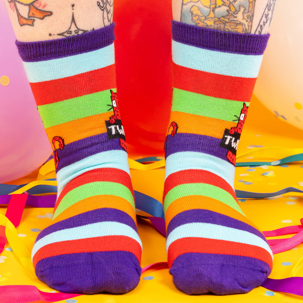A model wearing Katie Abey socks with a red cat holding a sign saying twat. They are striped in multi colours and lovely and vibrant. The model is stood on a yellow floor with balloons, confetti and ribbons