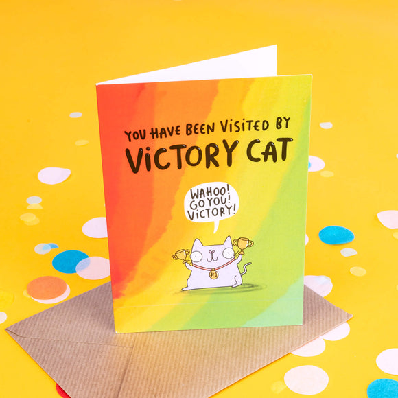 A rainbow coloured greeting card with black writing on front saying 'you have been visited by victory cat' and a grey cat illustration with red collar holding gold trophies and has a speech bubble above it's head that reads 'wahoo! go you! victory!'