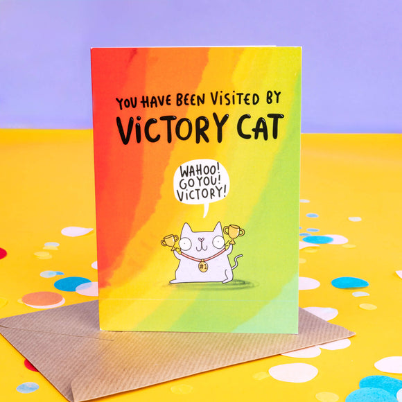 A rainbow coloured greeting card with black writing on front saying 'you have been visited by victory cat' and a grey cat illustration with red collar holding gold trophies and has a speech bubble above it's head that reads 'wahoo! go you! victory!'