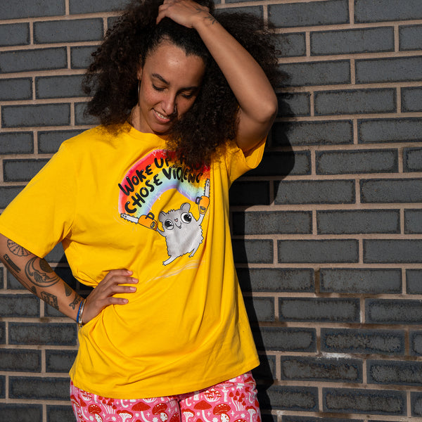 Person posing in the Katie Abey Woke Up and Chose Violence Hamster Adults Tee in Spectra Yellow. The yellow unisex t shirt features a colourful hamster illustration holding chainsaws with a rainbow slogan. Designed by UK illustrator Katie Abey, this quirky adult tee is ideal for bold, playful and alternative wardrobes.
