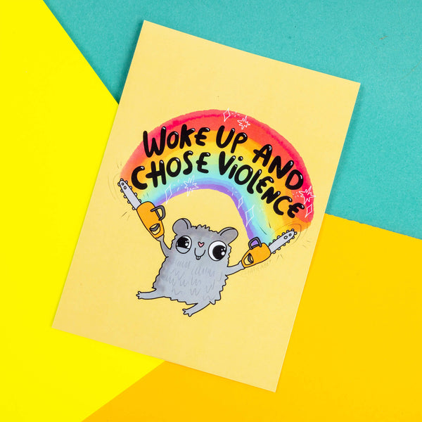 A yellow postcard of a hamster with two chainsaws and a rainbow above its head with text saying woke up and chose violence illustrated by Katie Abey