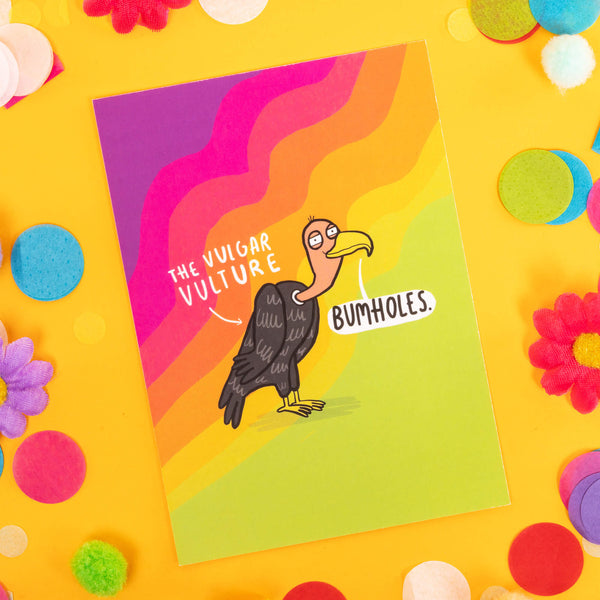 a postcard of an illustration drawn by Katie Abey of the Vulgar Vulture saying 'Bumholes' against a rainbow wavy background. It is laid on a yellow backdrop with confetti, fake daisies and pom poms of various colours