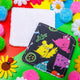 a dark grey square shaped makeup remover pad with colourful Katie Abey character illustrations on it. Next to it is another makeup remover pad sat face down to show the plain white side of the pad. They are laying on top of a bright yellow backdrop with coloured confetti and flowers scattered around
