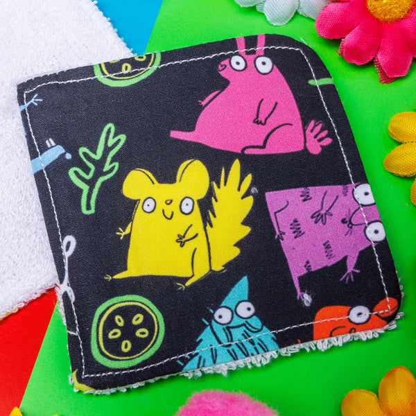 a dark grey square shaped makeup remover pad with colourful Katie Abey character illustrations on it. They are laying on top of a bright yellow backdrop with coloured confetti and flowers scattered around