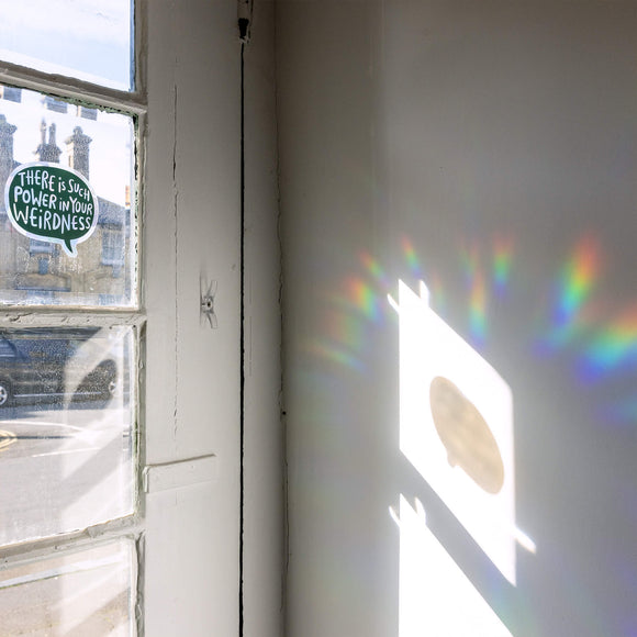 A window sticker shaped like a speech bubble stuck to a window that says there is such power in your weirdness. It is a black base and casts rainbows on a room. Designed By Katie Abey