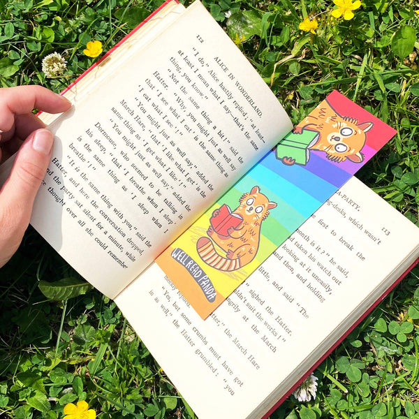 Well read panda bookmark laying face up in an open book laying on grass. The bookmark is laser printed card with a rainbow stripe background with two red pandas holding and reading books with bottom text reading 'Well Read Panda'. Designed by Katie Abey in the UK and printed with a silk finish so they are wipe clean.