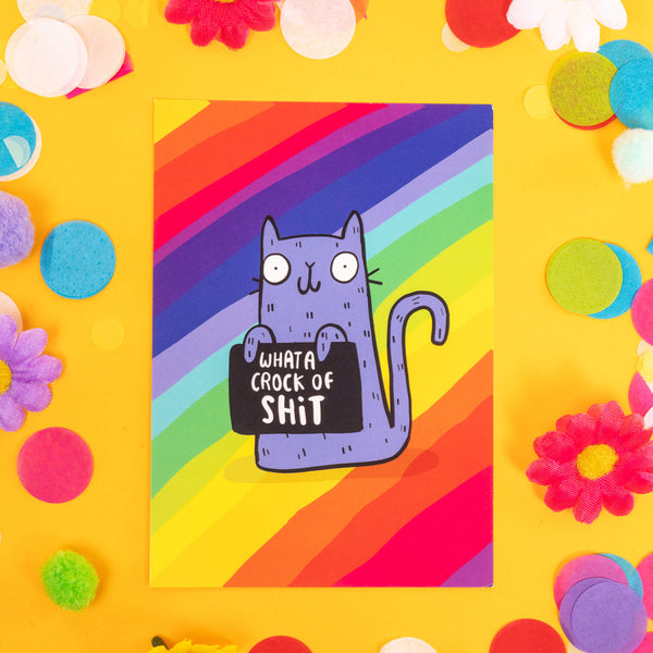 A postcard with a rainbow background and a purple cat holding a sign saying 'what a crock of shit' the card is laying on a yellow background with colourful confetti, fake daisies and pom poms 