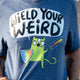 Close up of the Wield Your Weird Adults Tee in dark heather blue by Katie Abey. The front graphic features a quirky green cat holding a rainbow sword and shield with bold text reading Wield Your Weird. The illustration highlights Katie Abey’s playful art style and colourful design.
