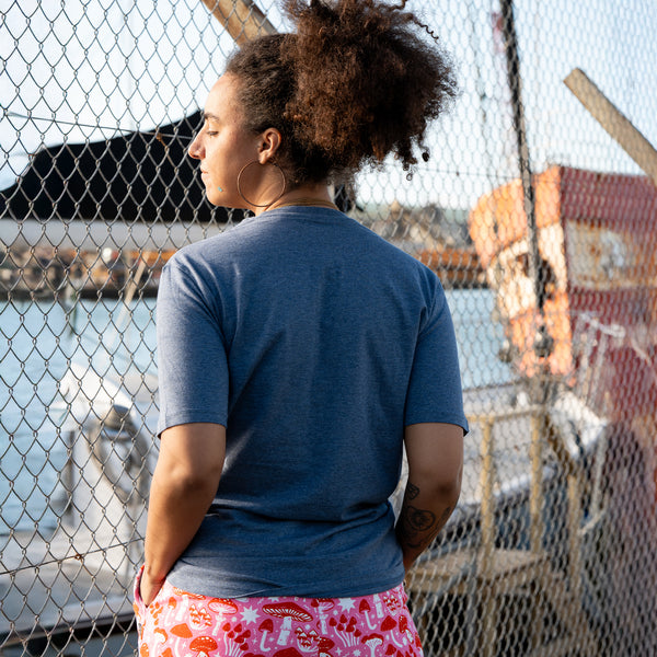 Back view of a model wearing the Wield Your Weird Adults Tee in dark heather blue by Katie Abey. The unisex t shirt has a relaxed fit and is styled casually outdoors by the water, showing the plain back and soft fabric drape. Ideal for showcasing fit and length from behind.
