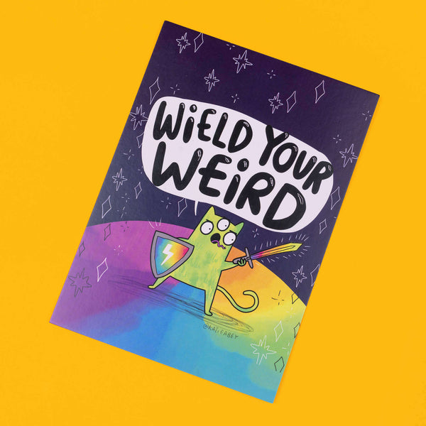 Colourful postcard by Katie Abey with the phrase "Wield Your Weird" in bold black letters on a white speech bubble. Below, a green, three-eyed cartoon cat stands on a rainbow ground, holding a shield and sword, ready for adventure. The background is a dark cosmic scene with stars and abstract shapes. The artist's handle, @katieabey, is visible. The background is bright yellow