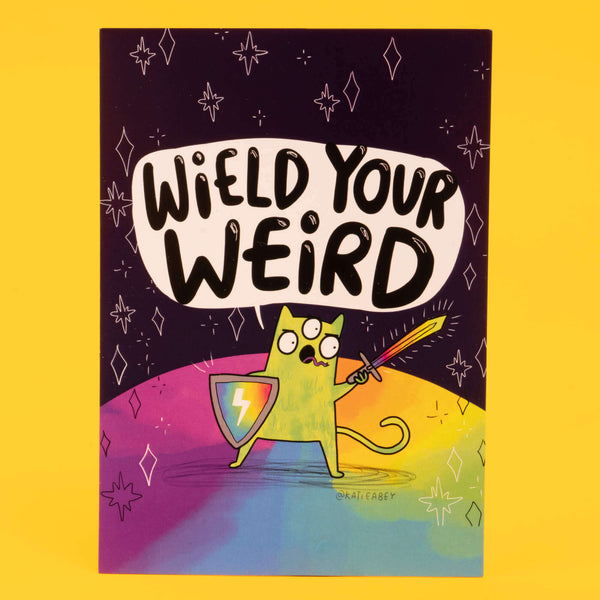 Colourful postcard by Katie Abey with the phrase "Wield Your Weird" in bold black letters on a white speech bubble. Below, a green, three-eyed cartoon cat stands on a rainbow ground, holding a shield and sword, ready for adventure. The background is a dark cosmic scene with stars and abstract shapes. The artist's handle, @katieabey, is visible. The background is bright yellow