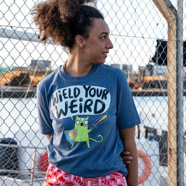 Three quarter view of a model wearing the Wield Your Weird Adults Tee in dark heather blue by Katie Abey. The playful slogan and illustrated cat design are clearly visible, styled casually outdoors. A comfortable adults graphic t shirt with a relaxed fit and positive message.
