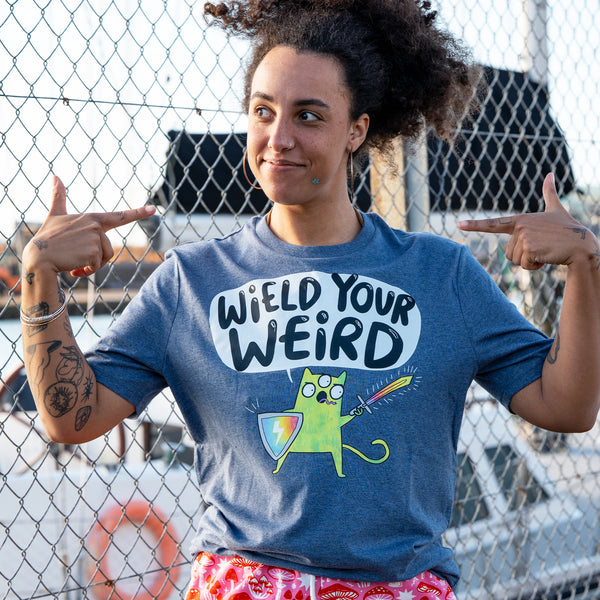 Front view of a model wearing the Wield Your Weird Adults Tee in dark heather blue by Katie Abey. The model points at the slogan on the t shirt, clearly showing the fun illustrated cat character and bold lettering. A joyful unisex graphic tee designed to celebrate being weird.
