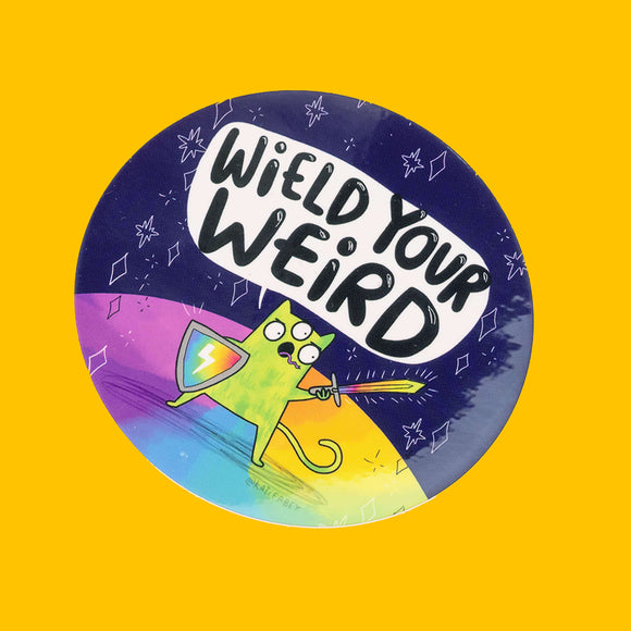 Colourful circular vinyl sticker by Katie Abey with the phrase "Wield Your Weird" in bold black letters on a white speech bubble. Below, a green, three-eyed cartoon cat stands on a rainbow ground, holding a shield and sword, ready for adventure. The background is a dark cosmic scene with stars and abstract shapes. The artist's handle, @katieabey, is visible. The background is bright yellow