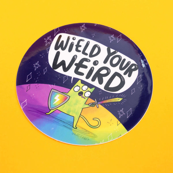 Colourful circular vinyl sticker by Katie Abey with the phrase "Wield Your Weird" in bold black letters on a white speech bubble. Below, a green, three-eyed cartoon cat stands on a rainbow ground, holding a shield and sword, ready for adventure. The background is a dark cosmic scene with stars and abstract shapes. The artist's handle, @katieabey, is visible. The background is bright yellow