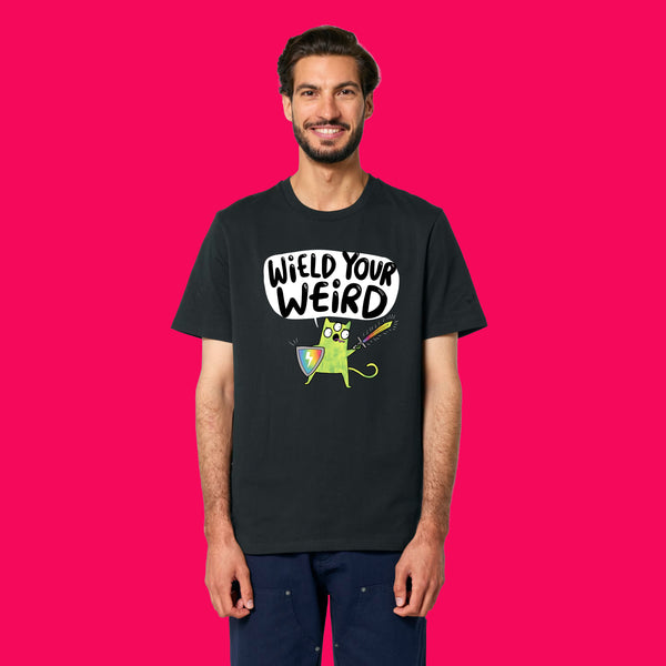 Front view of a model wearing the Wield Your Weird Adults T Shirt by Katie Abey in black. The playful illustrated slogan design is clearly visible on the chest, styled with dark trousers on a bright pink background.