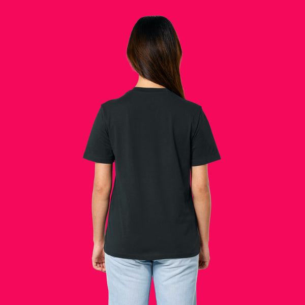 Back view of a model wearing the Wield Your Weird Adults T Shirt by Katie Abey in black. The relaxed fit short sleeve T shirt is styled with light blue jeans and photographed on a bright pink background.