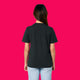 Back view of a model wearing the Wield Your Weird Adults T Shirt by Katie Abey in black. The relaxed fit short sleeve T shirt is styled with light blue jeans and photographed on a bright pink background.