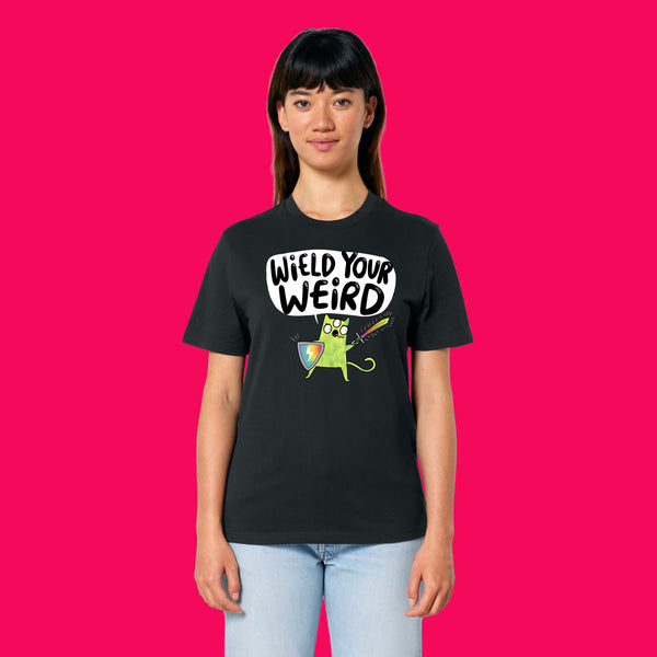 Front view of a model wearing the Wield Your Weird Adults T Shirt by Katie Abey in black. The bold illustrated character and slogan are visible on the chest, paired with light blue jeans and photographed against a bright pink background.