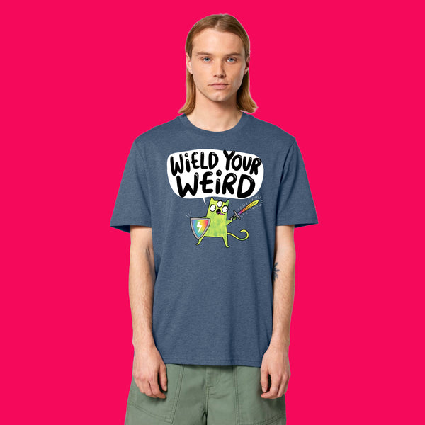 Front view of a model wearing the Wield Your Weird Adults T Shirt by Katie Abey in dark heather blue. The bold illustrated slogan design is clearly visible on the chest, paired with green trousers and photographed against a bright pink background.