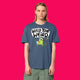 Front view of a model wearing the Wield Your Weird Adults T Shirt by Katie Abey in dark heather blue. The bold illustrated slogan design is clearly visible on the chest, paired with green trousers and photographed against a bright pink background.