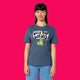 Front view of a model wearing the Wield Your Weird Adults T Shirt by Katie Abey in dark heather blue. The playful illustrated slogan design is visible on the chest, paired with green trousers and photographed on a bright pink background.