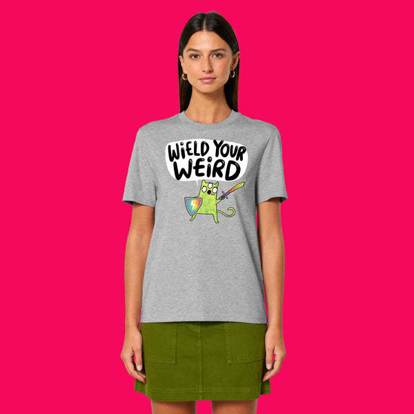 Front view of a model wearing the Wield Your Weird Adults T Shirt by Katie Abey in heather grey. The T shirt features a quirky illustrated character with a sword and shield and the slogan Wield Your Weird, styled with a green skirt on a bright pink background.