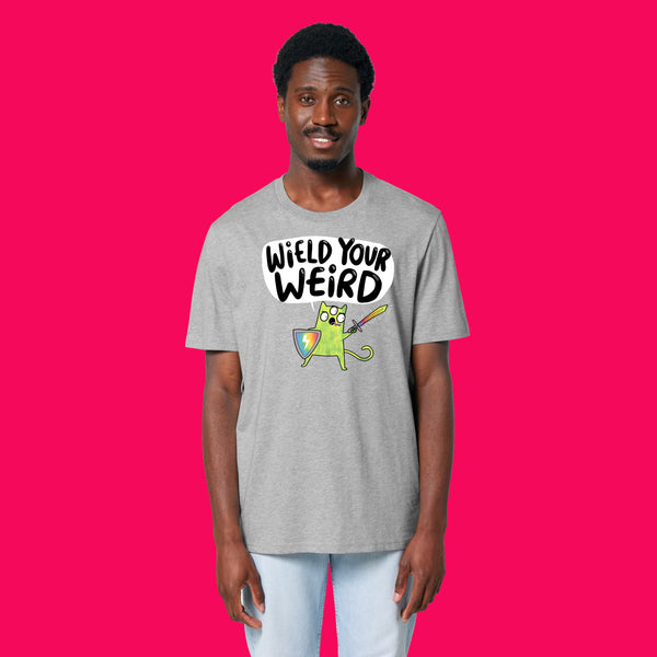 Front view of a model wearing the Wield Your Weird Adults T Shirt by Katie Abey in heather grey. The illustrated slogan design is clearly visible on the chest, paired with light blue jeans and photographed on a bright pink background.