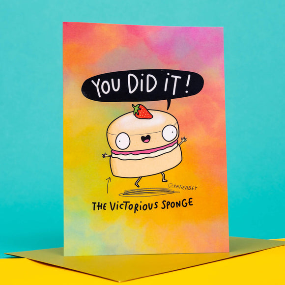 funny congratulations A6 card featuring The Victorious Sponge smiling strawberry on it's head and speech bubble that reads 'You did it!' on a rainbow coloured card with pairing gold envelope. Designed by Katie Abey.