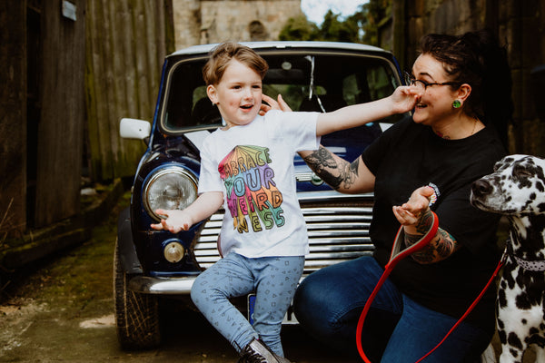 Toddlers Kids childrens white cotton T-shirt reading 'Embrace Your Weirdness' printed in bold large colourful rainbow letters on the front, and small on the front right hand side, in red orange yellow green teal blue purple and pink. Designed by Katie Abey in the UK.