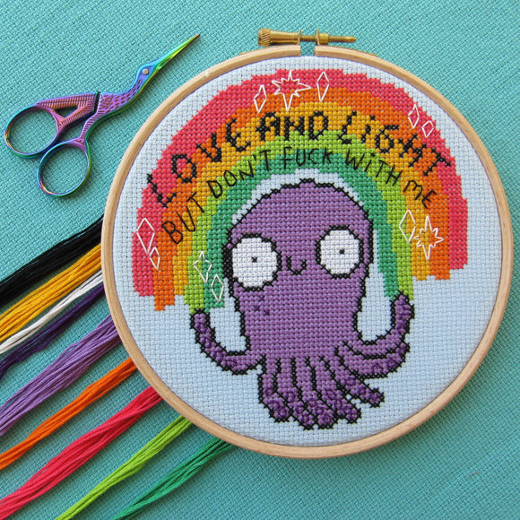 Love and Light octopus cross stitch kits, a collaboration between hoops and crosses and katie abey illustrations. An embroidery hoop is sat on teal fabric with multicoloured embroidery threads and scissors surrounding it. The cross stitch design in the embroidery hoop is a rainbow sitting above a purple octopus with white sparkles and black text that reads 'Love and Light but don't fuck with me'.