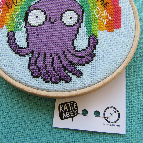 Love and Light octopus cross stitch kits, a collaboration between hoops and crosses and katie abey illustrations. A close up on an embroidery hoop sat on teal fabric with a business card underneath. The cross stitch design in the embroidery hoop is a rainbow sitting above a purple octopus with white sparkles and black text that reads 'Love and Light but don't fuck with me'.