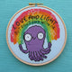 Love and Light octopus cross stitch kits, a collaboration between hoops and crosses and katie abey illustrations. An embroidery hoop is sat on teal fabric. The cross stitch design in the embroidery hoop is a rainbow sitting above a purple octopus with white sparkles and black text that reads 'Love and Light but don't fuck with me'.