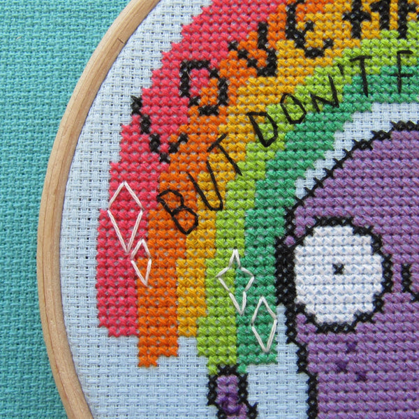 Love and Light octopus cross stitch kits, a collaboration between hoops and crosses and katie abey illustrations. A close up on an embroidery hoop sat on teal fabric. The cross stitch design in the embroidery hoop is a rainbow sitting above a purple octopus with white sparkles and black text that reads 'Love and Light but don't fuck with me'.