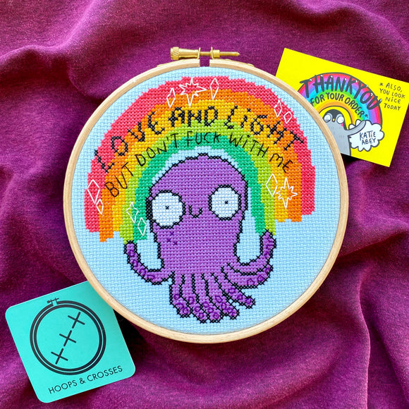 Love and Light octopus cross stitch kits, a collaboration between hoops and crosses and katie abey illustrations. An embroidery hoop is sat on purple fabric with two business cards surrounding it. The cross stitch design in the embroidery hoop is a rainbow sitting above a purple octopus with white sparkles and black text that reads 'Love and Light but don't fuck with me'.