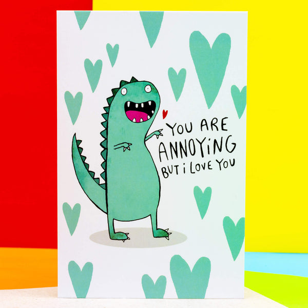 You're annoying but I love you a6 greeting card designed and printed by Katie Abey in the UK. The card is stood upright on an orange background. The card front cover is a white background with a green t-rex style dinosaur smiling with multiple green hearts surrounding it and text reading 'You are annoying but I love you'.
