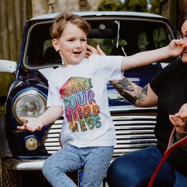 Toddlers Kids childrens white cotton T-shirt reading 'Embrace Your Weirdness' printed in bold large colourful rainbow letters on the front, and small on the front right hand side, in red orange yellow green teal blue purple and pink. Designed by Katie Abey in the UK.