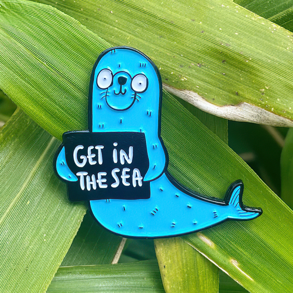 'Get In The Sea' soft enamel pin featuring smiley happy fun blue seal with black outline, holding black sign with white lettering. 35mm with rubber clasp. Designed by Katie Abey in the UK
